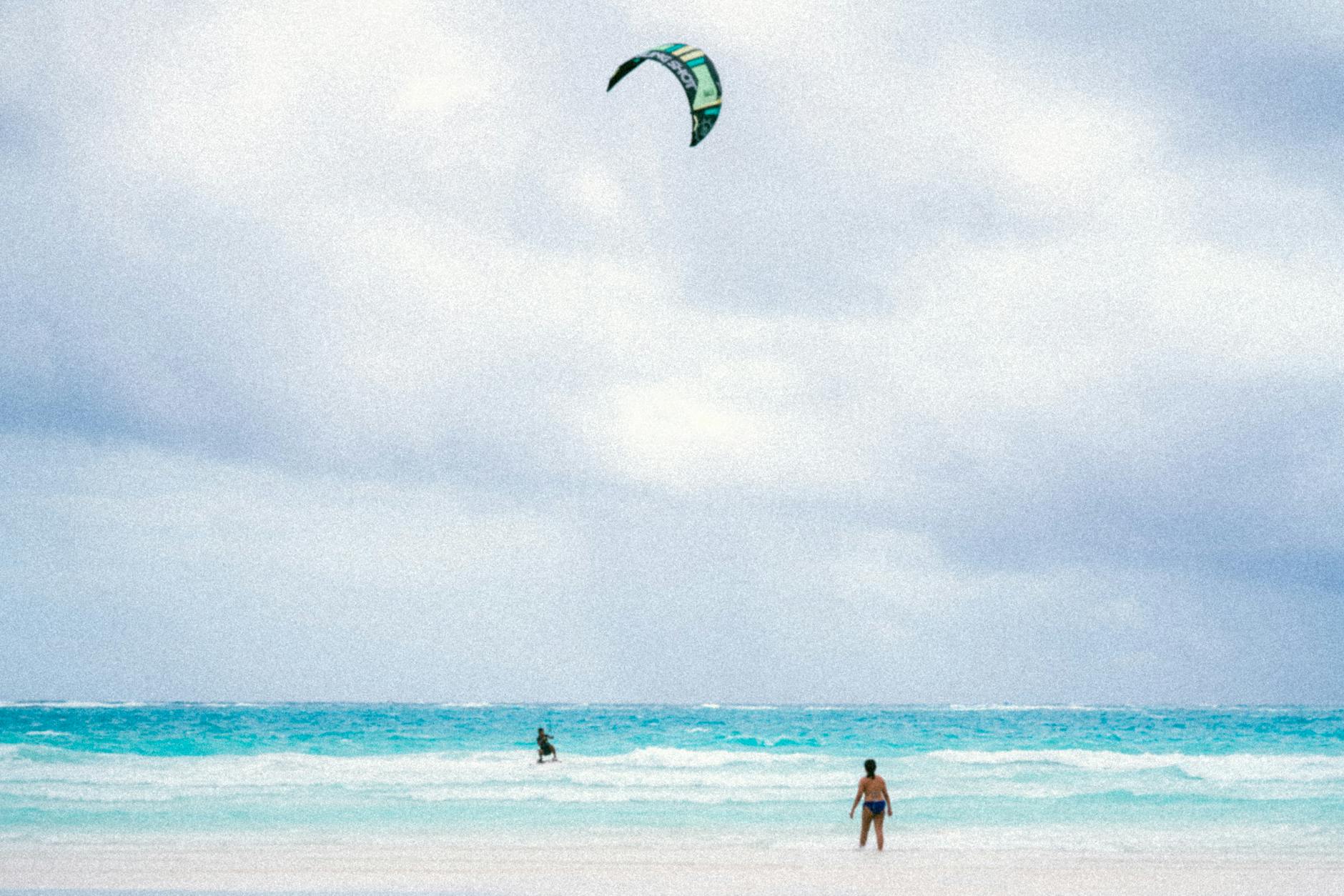 A person enjoying kitesurfing on a serene tropical beach with clear blue waters and cloudy skies.