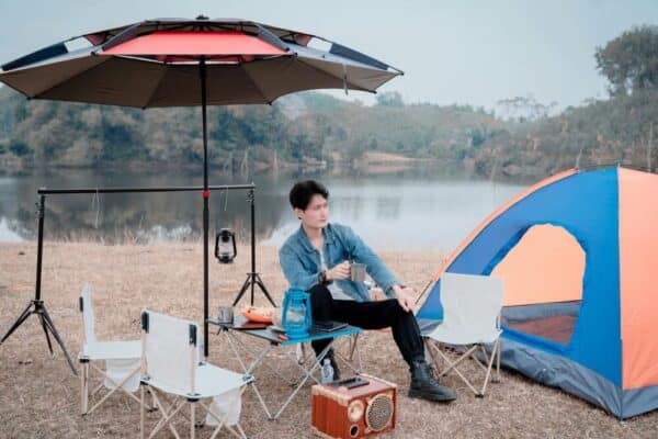 戶外露營場景，青年男子在湖邊搭建帳篷，享受自然風光，配備帳篷、折疊椅、露營燈與便攜音箱，展現現代露營休閒生活方式。.