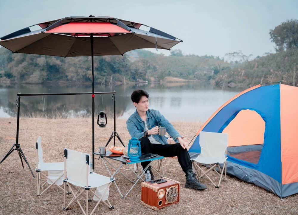 戶外露營場景，青年男子在湖邊搭建帳篷，享受自然風光，配備帳篷、折疊椅、露營燈與便攜音箱，展現現代露營休閒生活方式。.
