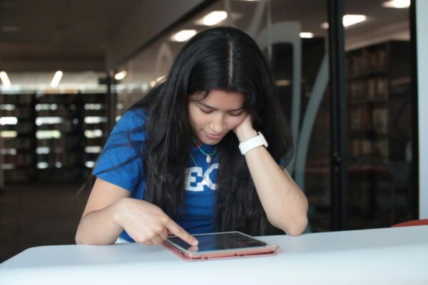 A young woman in a blue t-shirt is using a tablet at a library table. Focused and engaged in her task.