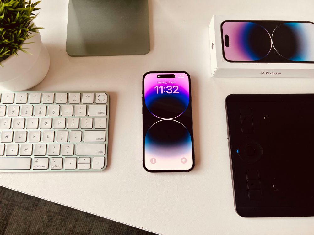 A sleek workspace setup featuring an iPhone with colorful display beside a keyboard and plant.