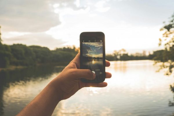 Hand photographing peaceful lake scenery with a smartphone at sunset.