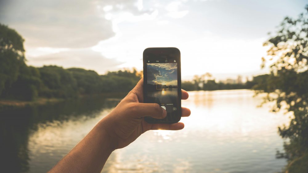 Hand photographing peaceful lake scenery with a smartphone at sunset.