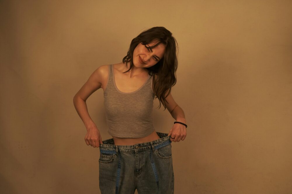 A woman smiles while holding out the waistband of oversized jeans to show her weight loss success.