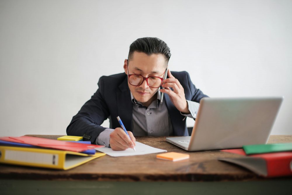 Professional businessman multitasking with phone call and laptop while writing notes at desk.