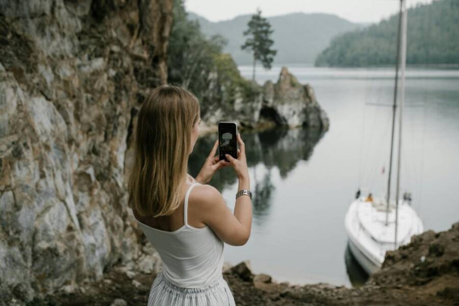 A woman takes a photo of a serene lakeside with a sailboat in lush natural surroundings.