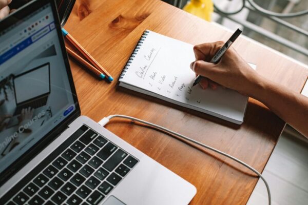 打造常銷型線上課程完整流程:Evergreen Course 一步步指南 A person writing notes while researching online courses on a laptop in a home setting.