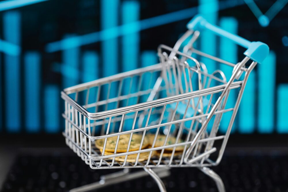 A miniature shopping cart filled with coins against a digital financial graph background.