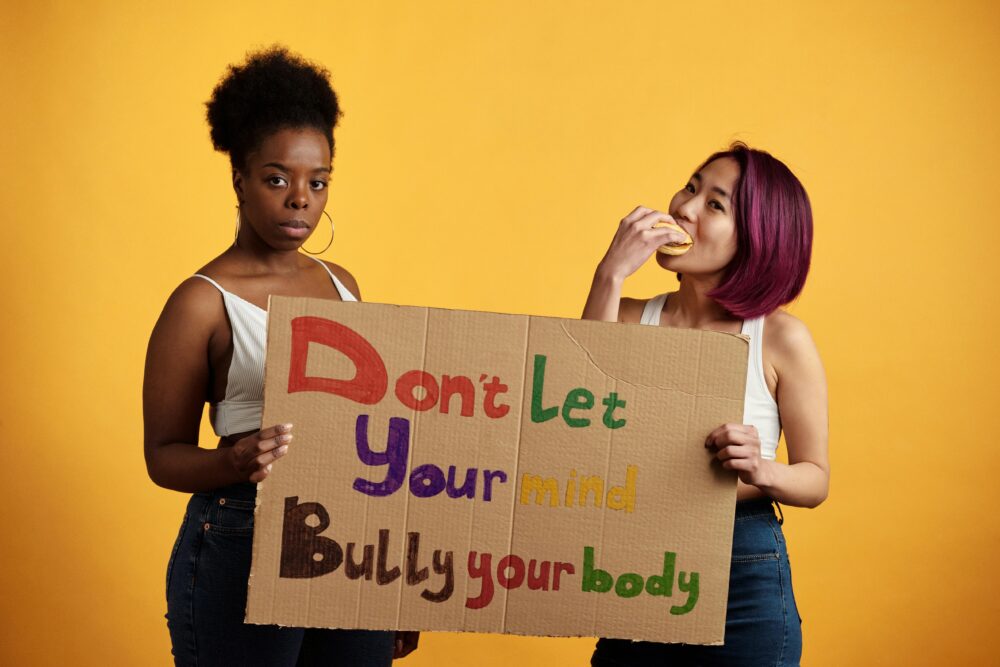 Two diverse women with empowering message against body shaming on a yellow background.