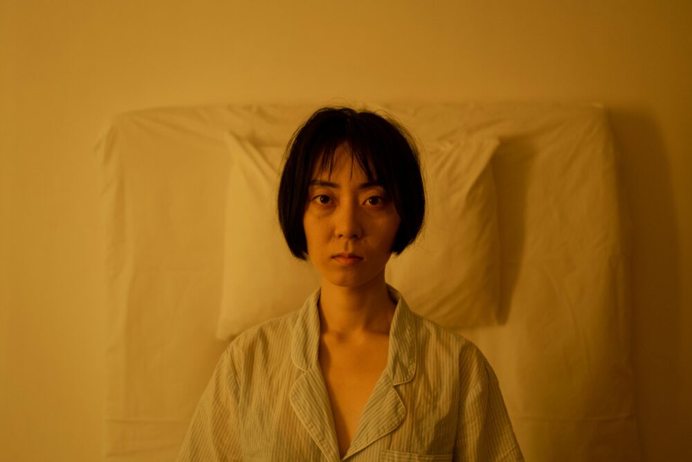 An Asian woman sits in bed with an expression of insomnia and stress, enveloped in a yellow ambiance.