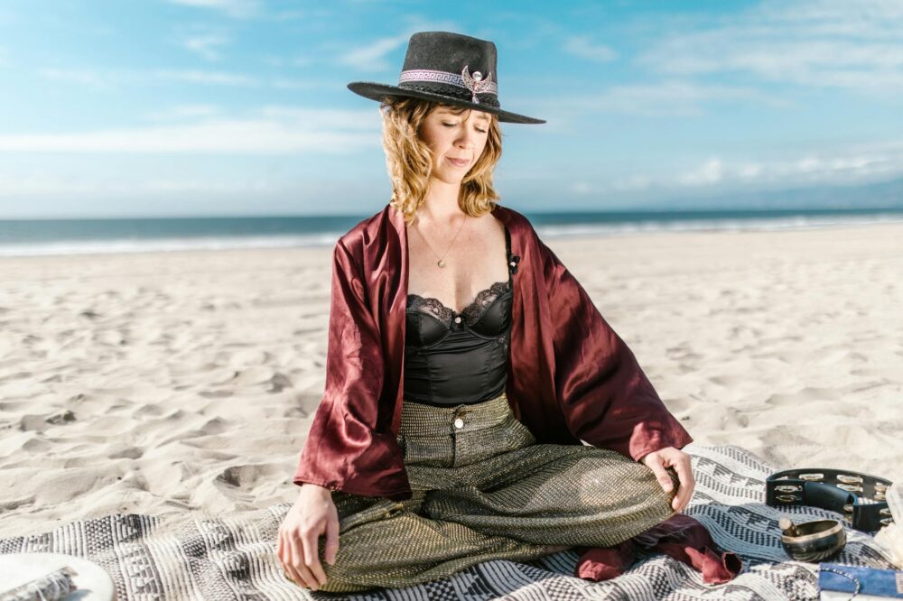 Stylish woman meditating on a beach in elegant attire with a serene backdrop.