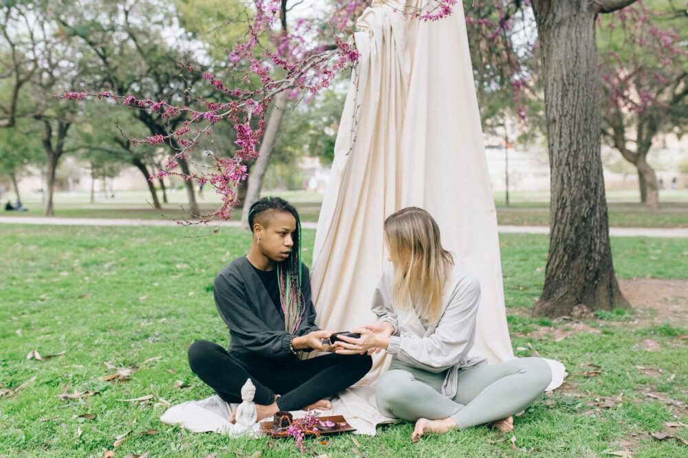 Two women engage in a peaceful meditation session outdoors under blossoming trees, promoting tranquility and connection.
