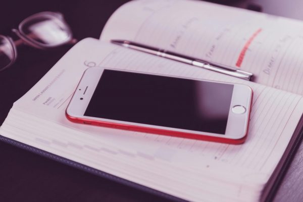 行事曆備忘錄跨系統遷移指南:手機資料輕鬆轉移 A red and white smartphone on an open diary accompanied by glasses and a pen.