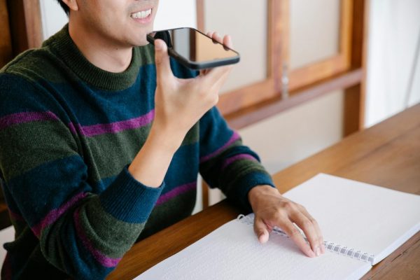 Adult man using smartphone for voice commands, smiling at home desk.