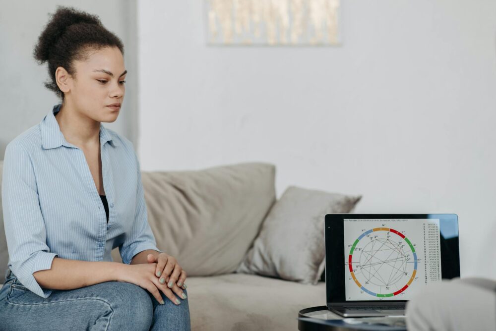 Young woman sitting in a living room analyzing an astrology chart on a laptop, reflecting concentration.