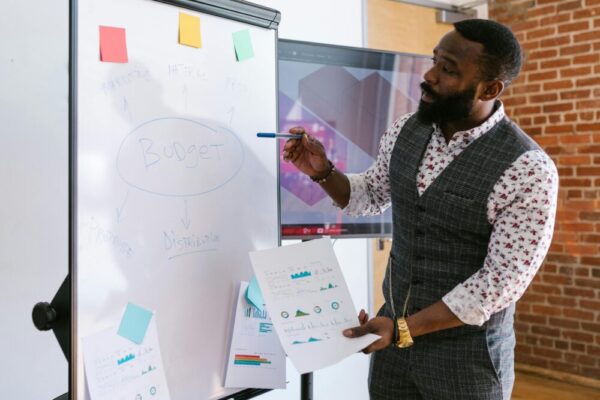 A businessman explains budget strategies using a whiteboard in an office setting.