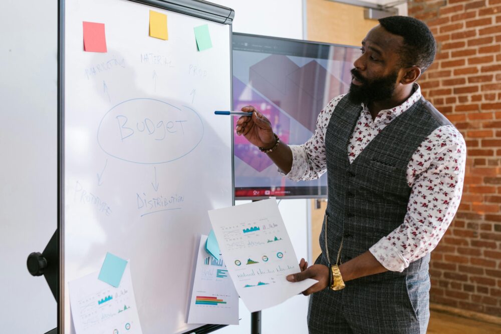A businessman explains budget strategies using a whiteboard in an office setting.