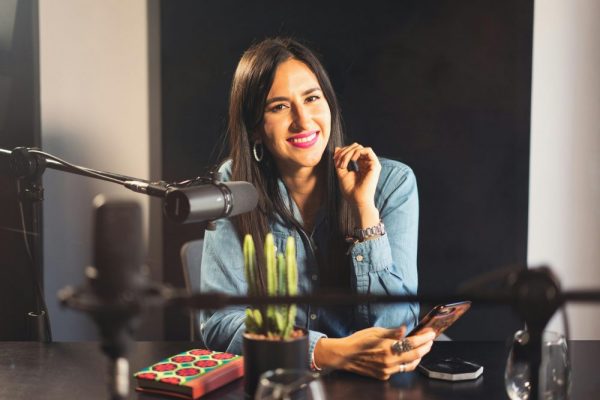 Smiling woman in denim recording a podcast with microphone, phone, and cactus on table.