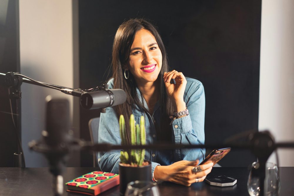 Smiling woman in denim recording a podcast with microphone, phone, and cactus on table.