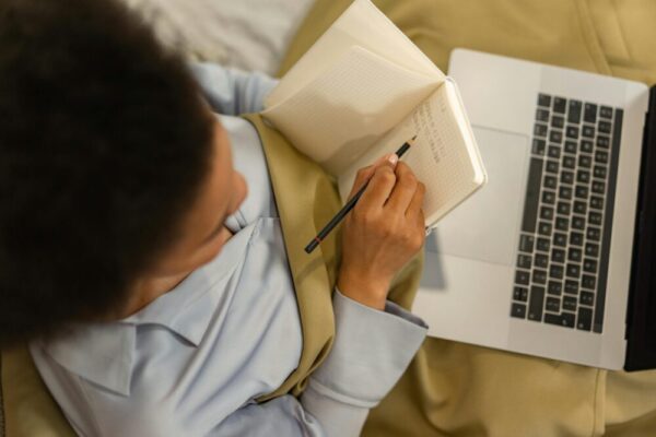 線上課程平台比較:Teachable、Thinkific、Kajabi 該選哪個? Overhead view of a woman taking notes in a notebook while using a laptop, embodying home learning.