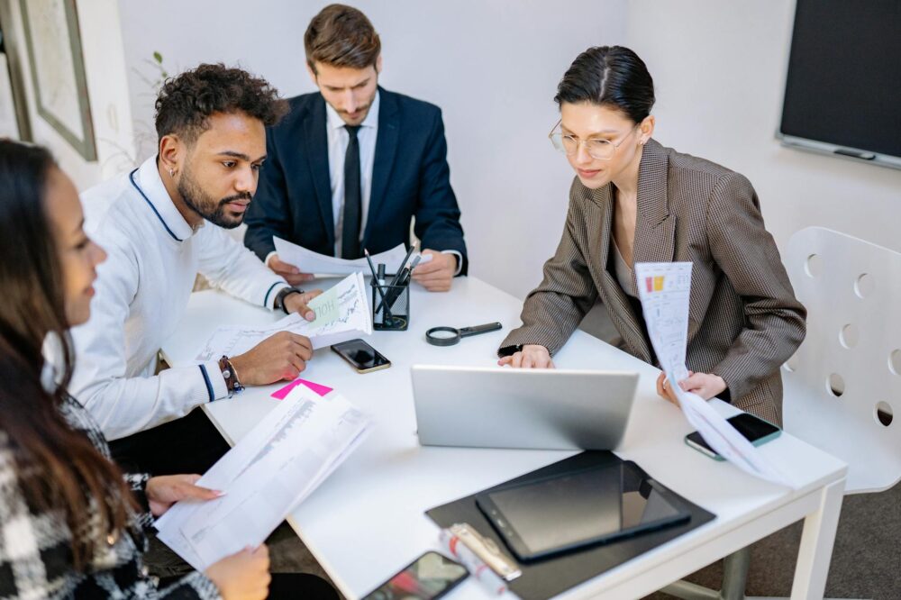 A diverse group of professionals in a business meeting, collaborating over documents and digital devices.