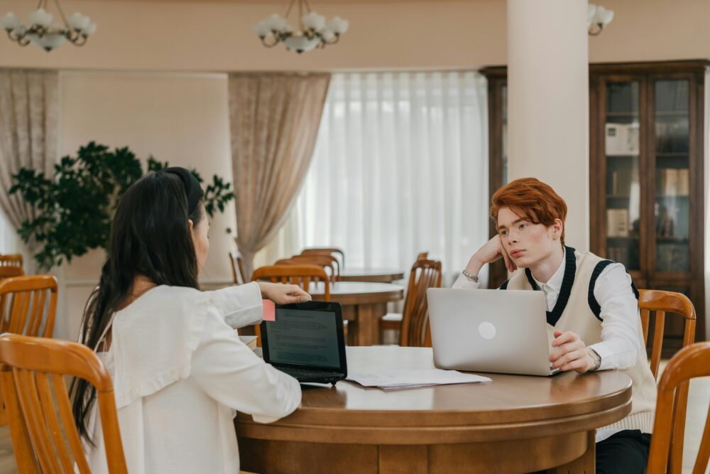 Two students using laptops for study in a warm, indoor library setting.