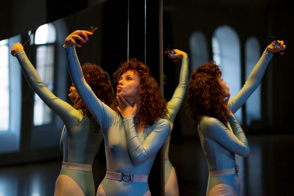 Young woman in leotard takes a selfie with reflection in a dimly lit room, showcasing a modern indoor setting.