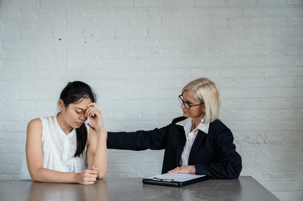 A supportive counseling session where one woman comforts another at a table against a brick wall.
