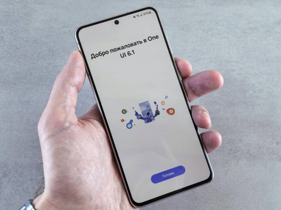 A close-up of a hand holding a smartphone displaying One UI 6.1 setup screen with Cyrillic text
