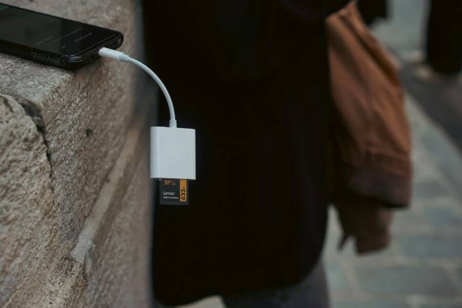 A mobile phone connected to a memory card reader outdoors on a stone surface.