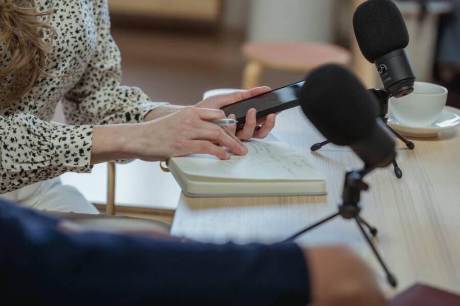 Woman taking notes and using smartphone on a podcast recording setup indoors.