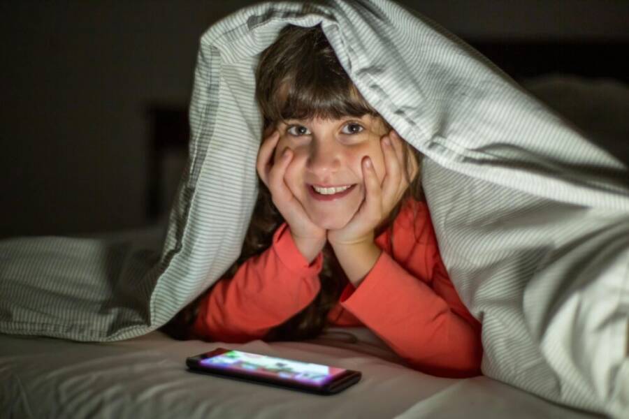 A young girl smiling under a blanket, using a smartphone at night indoors
