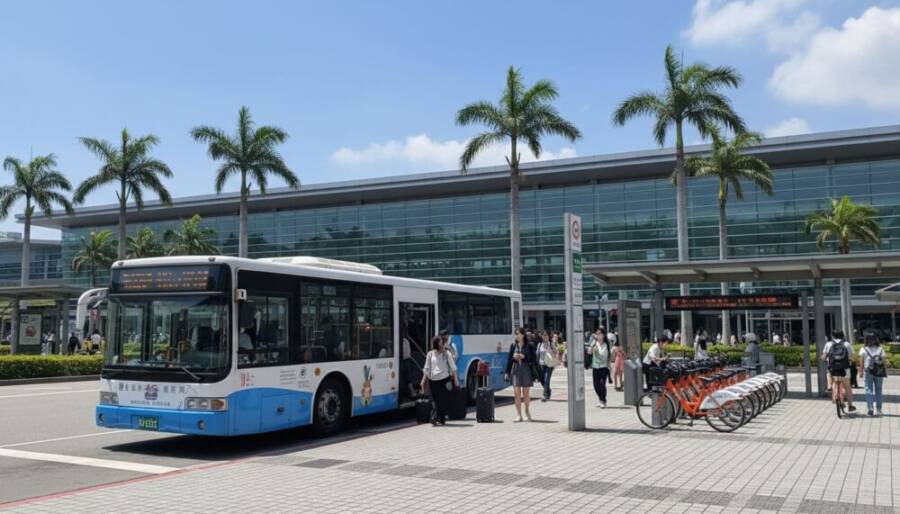 A vibrant scene at Taichung HSR station in Taiwan, showing people boarding a public bus and others renting iBike shared bicycles nearby.