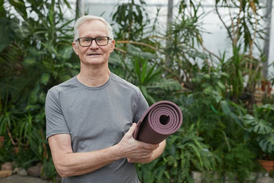 Smiling senior man with gray hair holding a yoga mat in a lush greenhouse